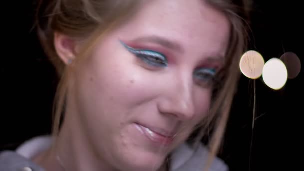 Retrato de menina loira com maquiagem colorida tentando soprar o cabelo do rosto em luzes borradas fundo . — Vídeo de Stock