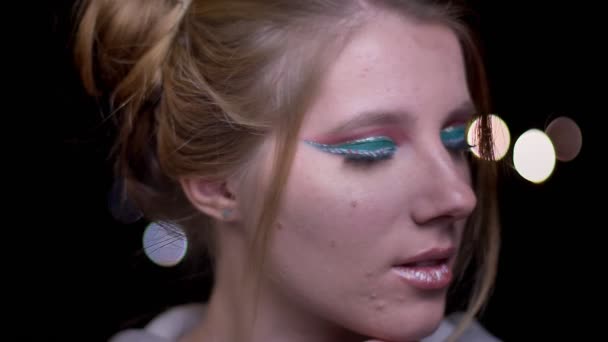 Close-up portret van blonde model met kleurrijke make-up fashionably poseren in camera op wazig lights achtergrond. — Stockvideo