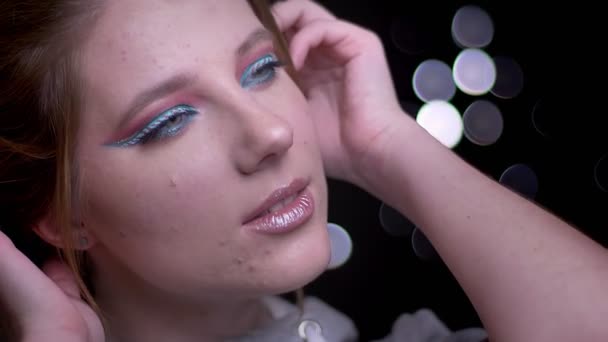 Blond modell med färgglada make-up trendigt poserar i kameran och vinden blåser hennes hår på suddig ljus bakgrund. — Stockvideo