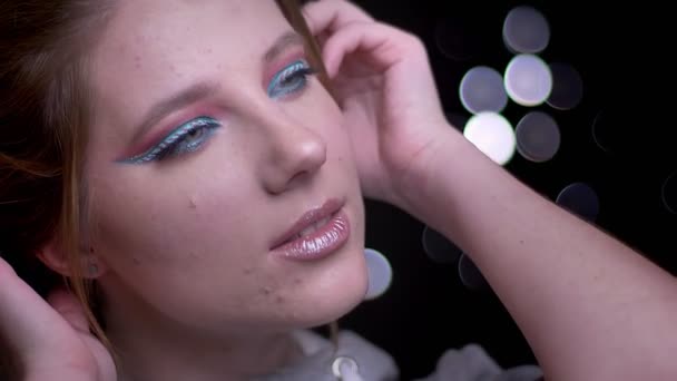 Modello biondo con trucco colorato alla moda in posa in macchina fotografica e vento soffiando i capelli su sfondo luci sfocate . — Video Stock