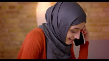 Closeup çekim genç çekici Müslüman kadın çalışanın raslantı telefonda konuşurken ve neşeyle kapalı ofis içinde belgili tanımlık laptop önünde oturan gülümseyen Hicap