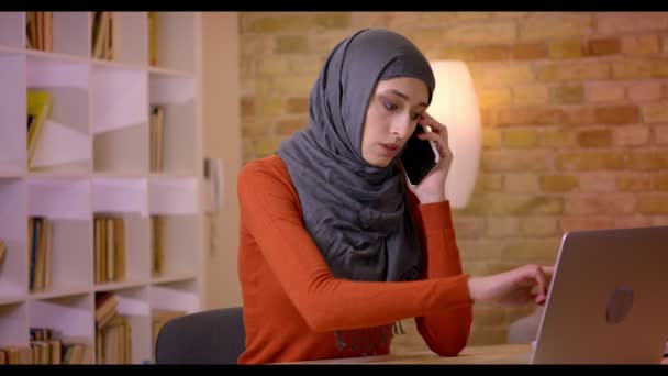 Närbild-shoot av unga attraktiva muslimsk kvinnlig anställd i hijab har en konversation på telefonen medan du skriver på den bärbara datorn inomhus på kontoret — Stockvideo