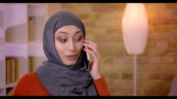 Primo piano di tiro di giovane impiegata musulmana attraente in hijab avendo una telefonata e sorridendo allegramente seduto davanti al computer portatile all'interno dell'ufficio — Video Stock