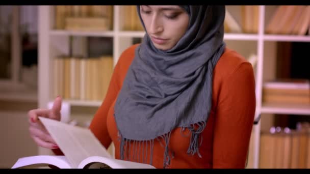 Tiro de close-up de jovem estudante muçulmano atraente em hijab lendo um livro em pé dentro da biblioteca — Vídeo de Stock