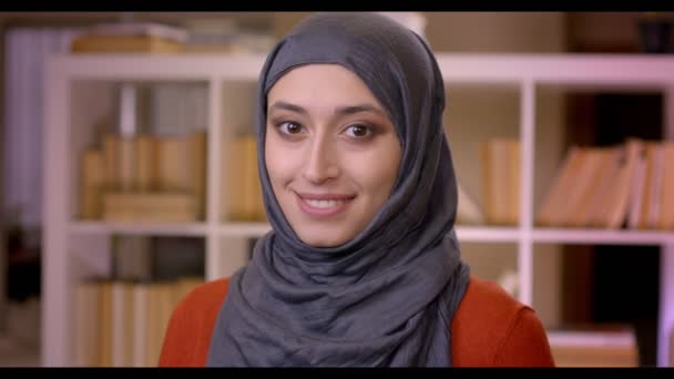 Closeup schieten van jonge aantrekkelijke moslim vrouwelijke student in hijab camera kijken en glimlachend gelukkig permanent binnenshuis in de bibliotheek — Stockvideo