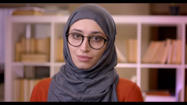 Zbliżenie strzelać z młodych atrakcyjnych muzułmańskich sukces kobiet student w hidżab i okulary, patrząc na aparat stały pomieszczeniu w bibliotece — Wideo stockowe