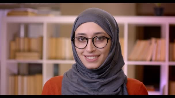 Sesión de primer plano de la joven atractiva estudiante árabe en hijab y gafas mirando a la cámara de pie en el interior de la biblioteca — Vídeo de stock