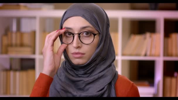 Gros plan de jeune étudiante musulmane séduisante en hijab fixant ses lunettes en regardant la caméra debout à l'intérieur de l'université de la bibliothèque — Video