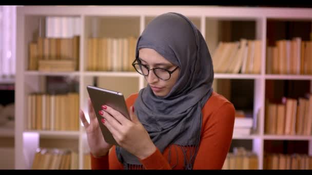 Primer plano de brote de joven atractiva estudiante musulmana en hijab escribiendo en la tableta de pie en el interior de la biblioteca — Vídeo de stock