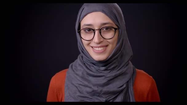 特写镜头拍摄年轻的有吸引力的穆斯林女性在头巾看着相机, 并愉快地微笑着与背景孤立在黑色 — 图库视频影像
