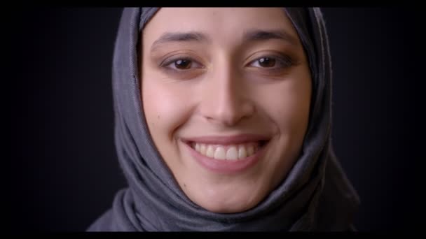 Sesión de primer plano de cara femenina musulmana atractiva joven en hijab mirando a la cámara con expresión facial sonriente con fondo aislado en negro — Vídeo de stock