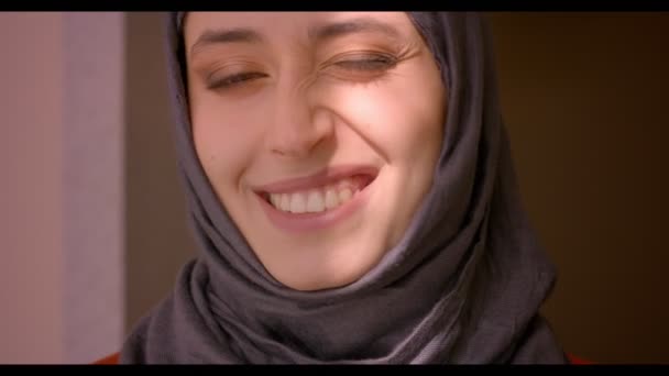 笑みを浮かべて、彼女の顔に輝く太陽と、目ウインクを自宅室内を作ってカメラを見て若い魅力的なイスラム教徒の女性のクローズ アップ撮影します。 — ストック動画