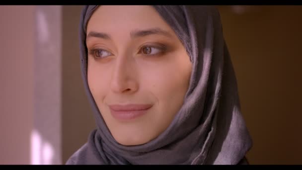 Syuting closeup dari perempuan muda muslim menarik menatap kamera dan tersenyum riang di dalam rumah dengan matahari bersinar di wajahnya — Stok Video