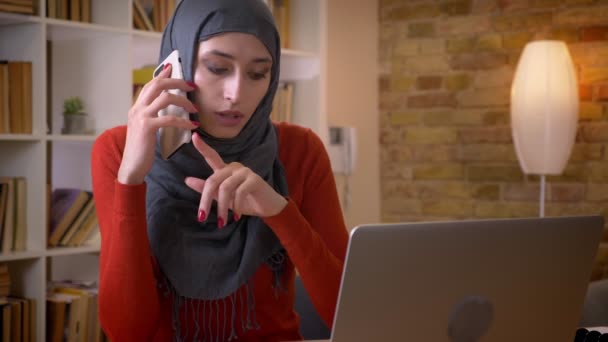 Närbild-shoot av unga attraktiva muslimska affärskvinna med ett telefonsamtal medan du skriver på den bärbara datorn inomhus på kontoret — Stockvideo