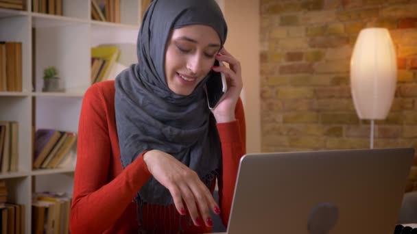 Primo piano sparare di giovane donna d'affari musulmana attraente in hijab avendo una conversazione telefonica durante la digitazione sul computer portatile all'interno dell'ufficio — Video Stock