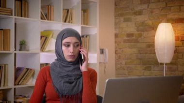 Closeup çekim genç çekici Müslüman işkadını kapalı ofis içinde belgili tanımlık laptop önünde oturan bir casualphone telefon sahip hijab içinde