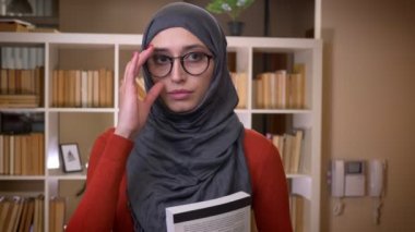 Öğrencinin genç çekici Müslüman kadın gözlüklerini sabitleme ve kapalı duran ve Kütüphane Üniversitesi'nde kamera düz bakarak bir kitap hijab portre çekimi