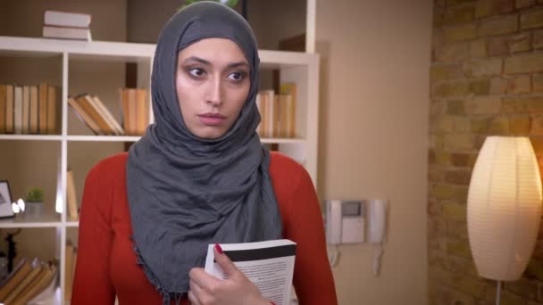 Closeup schieten van jonge aantrekkelijke moslim vrouwelijke student in hijab binnenshuis houden van een boek staan in de bibliotheek van de Universiteit — Stockvideo