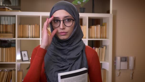 特写镜头拍摄年轻的有吸引力的穆斯林女学生在头巾固定她的眼镜, 拿着一本书站在室内, 直视相机在图书馆大学 — 图库视频影像