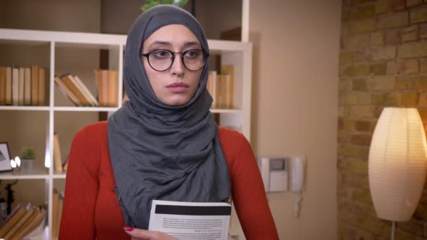 Closeup schieten van jonge aantrekkelijke moslim vrouwelijke student in hijab en glazen houden een boek staande binnenshuis in de bibliotheek van de Universiteit — Stockvideo