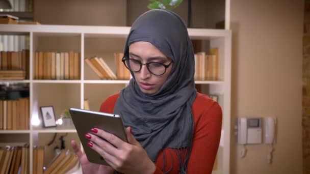 Primer plano de brote de joven atractiva estudiante musulmana en hijab navegar en la tableta de pie en el interior de la universidad de la biblioteca — Vídeos de Stock
