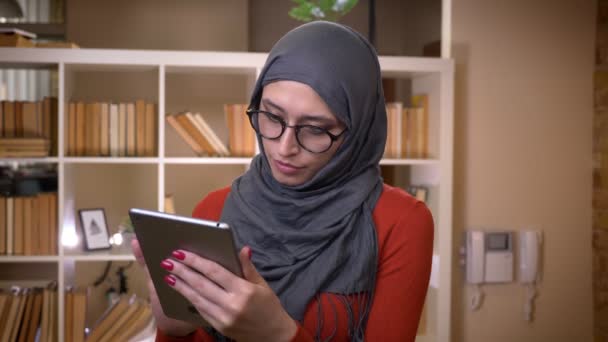 Sesión de primer plano de joven atractiva estudiante musulmana en hijab navegando en la tableta mirando hacia adelante y sonriendo felizmente de pie en el interior de la universidad de la biblioteca — Vídeos de Stock