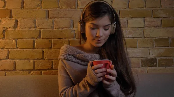 Portrait rapproché de jeunes femmes caucasiennes attrayantes écoutant de la musique dans des écouteurs assis sur le canapé et tenant une tasse de thé chaud — Photo