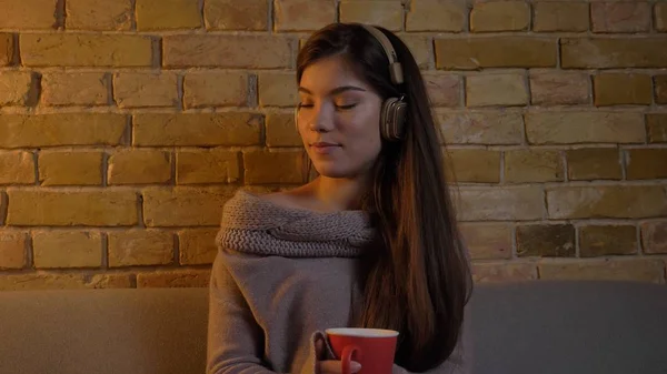 Portrait rapproché d'une jeune femme caucasienne écoutant de la musique dans un casque assis sur le canapé et dégustant une tasse de thé chaud dans un appartement confortable — Photo