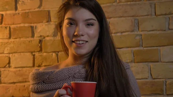 Närbild-shoot av unga attraktiva kaukasisk kvinna håller en kopp varmt te att vara glad och avslappnad titta på kameran och ler i en mysig lägenhet inomhus — Stockfoto