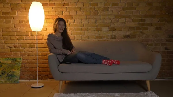 Closdeup σουτ του νέοι αρκετά καυκάσιος γυναίκα ακουμπά στον καναπέ και κρατώντας ένα τηλεχειριστήριο τηλεόρασης βλέποντας μια κωμωδία και πρόσχαρα γελώντας — Φωτογραφία Αρχείου