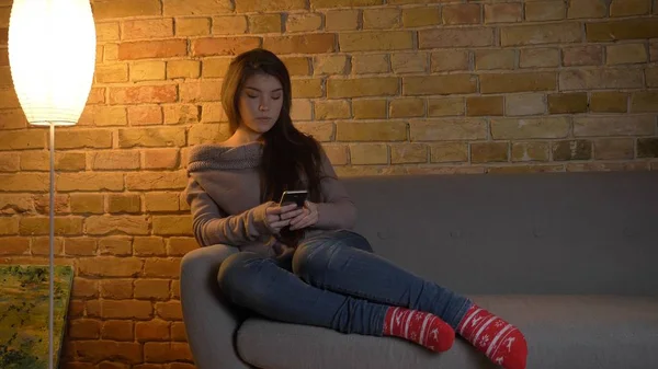 Nahaufnahme einer jungen hübschen kaukasischen Frau mit dem Telefon, während sie sich auf der Couch in einer gemütlichen Wohnung im Haus ausruht — Stockfoto