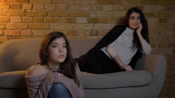 Retrato de close-up de duas jovens meninas bonitas assistindo TV com expressão facial curiosa enquanto descansa em um apartamento acolhedor dentro de casa — Fotografia de Stock