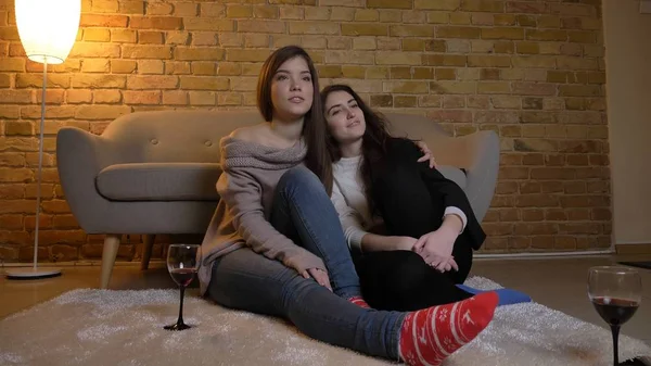 Closeup portrait of two young pretty women hugging and watching TV chilling with wine in a cozy apartment indoors