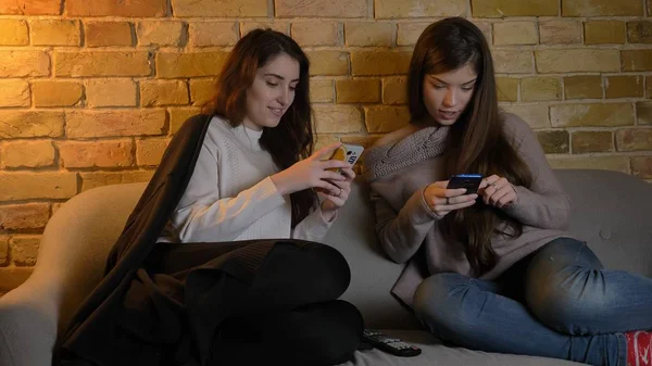 Крупный план портрета двух молодых симпатичных кавказских девушек, использующих телефоны, отдыхающих на диване в помещении — стоковое фото