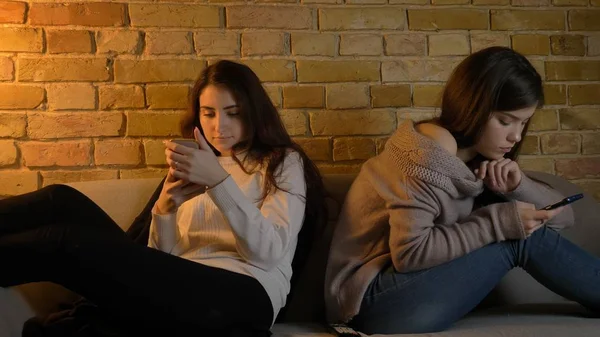 Крупный план портрета двух молодых симпатичных кавказских девушек, просматривающих телефоны, отдыхающих на диване в уютной квартире — стоковое фото