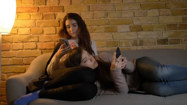 Retrato de cerca de dos chicas caucásicas jóvenes usando los teléfonos mientras descansan en el sofá en el interior. Uno tendido sobre los muslos de otro —  Fotos de Stock