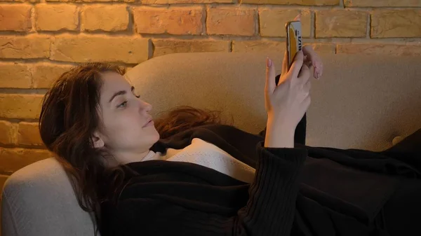 Schieten close-up van jonge mooie Kaukasische brunette vrouw liggend op de Bank en het gebruik van de telefoon binnenshuis in een gezellig appartement — Stockfoto