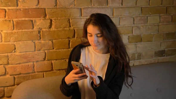 Close-up opnamen van jonge mooie Kaukasische brunette vrouwelijke zittend op de Bank en het gebruik van de telefoon binnenshuis in een gezellig appartement — Stockfoto