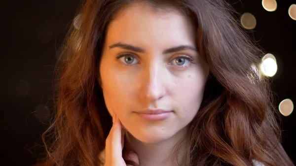 Nahaufnahme einer jungen hübschen kaukasischen Frau mit brünetten lockigen Haaren, die direkt in die Kamera mit Bokeh-Lichtern auf dem Hintergrund blickt — Stockfoto