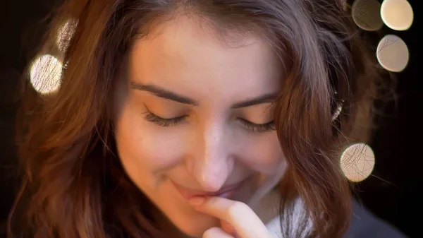 Close-up tiro de jovem mulher branca atraente sorrindo sedutoramente e mordendo o lábio na frente da câmera com luzes bokeh no fundo — Fotografia de Stock