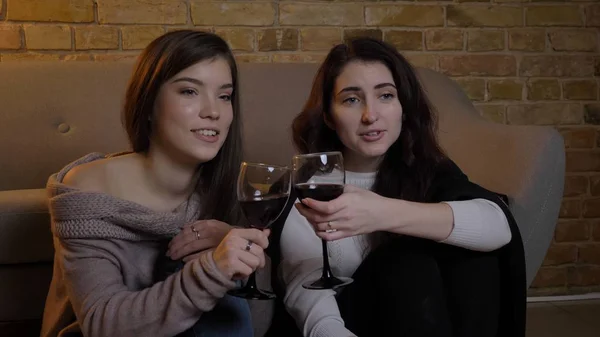 Nahaufnahme Porträt von zwei jungen hübschen Frauen, die in einer gemütlichen Wohnung im Haus mit Wein chillen und Sonnenbrillen klirren lizenzfreie Stockfotos