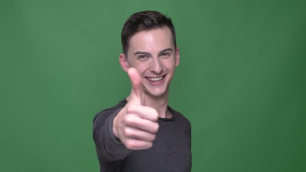 Closeup shoot of young handsome caucasian maleshowing thumb up smiling happily looking straight at camera with background isolated on green — Stock Video