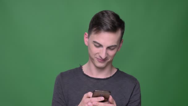 Nahaufnahme eines jungen gutaussehenden kaukasischen Mannes, der am Telefon SMS schreibt und in die Kamera lacht und lächelt, mit einem auf Grün isolierten Hintergrund — Stockvideo