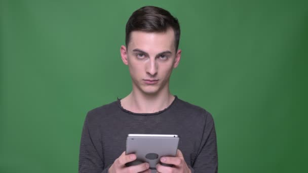 Primer plano de brote de joven guapo caucásico macho usando la tableta mirando a la cámara y sonriendo con fondo aislado en verde — Vídeos de Stock