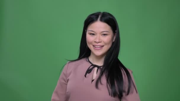 Closeup schieten van jonge aantrekkelijke Aziatische vrouw met zwarte haren glimlachen en gebaren duim omhoog het tonen van overeenkomst met geïsoleerd op groene achtergrond — Stockvideo