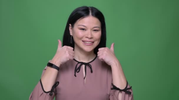 Closeup střílet z mladé atraktivní asijské ženy s černými vlasy, usmíval se a ukázal palcem nahoru říká Ano s pozadím izolované na zelené — Stock video