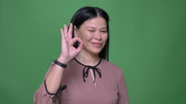 Närbild skjuta av unga attraktiva asiatisk kvinna med svart hår gestikulerande en okej handsign titta rakt på kameran med bakgrunden isolerad på grön — Stockvideo