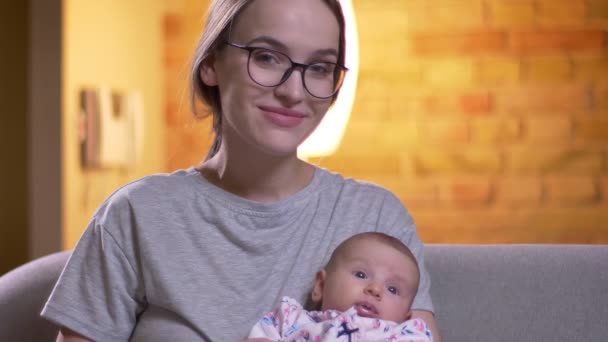 Nahaufnahme Porträt einer Mutter, die sich mit ihrer süßen neugeborenen Tochter umarmt und im Wohnzimmer ernsthaft in die Kamera schaut. — Stockvideo