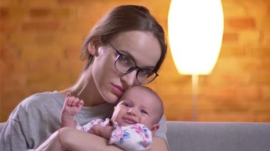 Yakın çekim portre ağlayan yeni doğan kızını tutan ve sakin bir şekilde oturma odasında kamera içine izlerken anne.