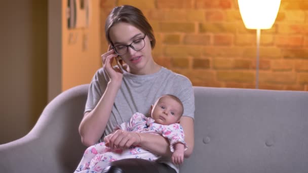 Nahaufnahme Porträt einer Mutter, die mit dem Smartphone spricht und ihre süße Tochter im Wohnzimmer hält. — Stockvideo